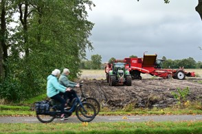 Voorbijgaand stel fietsers op fietspad kijken naar landbouwmachines die aan het aardappelen rooien zijn.