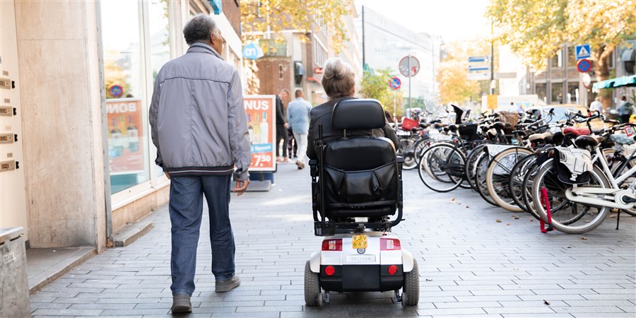 een ouder stel waarvan de vrouw in een scootmobiel zit die over de straat lopen