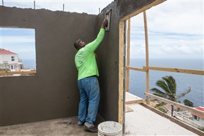 Man stuukt  wand van woning met op de achtergrond de zee en een palmboom