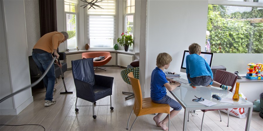 Een alleenstaande vader stofzuigt de huiskamer terwijl zijn beide zoons op een iMac computer spelletjes spelen