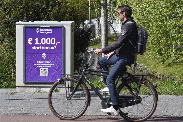 Jongen op fiets rijd langs een reclamebord met daar op de advertentie "Krantenjongens gezocht"