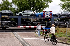 Een trein vervoert nieuwe auto&#x27;s en kruist een spoorwegovergang waar mensen voor de spoorbomen staan te wachten.