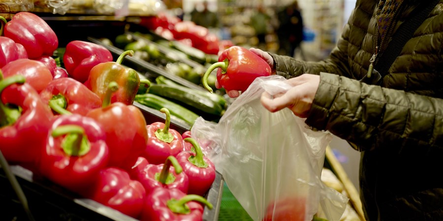 Een hand van een vrouw doet twee paprika's in een plasticzakje in de supermarkt.