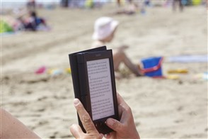 Op voorgrond leest iemand een e-reader, op achtergrond strand met spelend kind