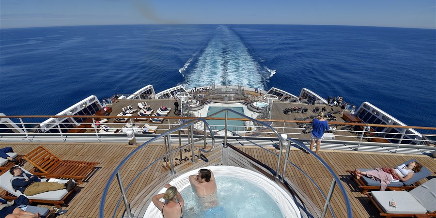 Passagiers liggen op zonnebedden op het achterdek van een varend cruiseschip. Twee passagiers genieten vanuit het zwembad van het uitzicht.