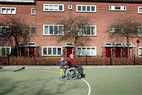 kleinzoon op inline skates achter de roelstoel van opa op sportveld in woonwijk