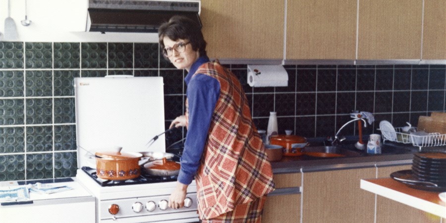 Vrouw in 70-er jaren broekpak aan het koken op gasfornuis.