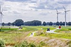 Landschap in Flevoland met windmolens, slingerende weg en naastgelegen fietspad met fietser, vrachtwagen, tractor en boeren met landbouwmachines op de akkers bezig met de oogst