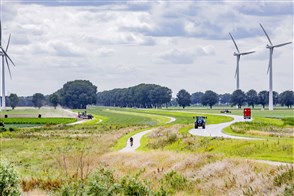 Landschap in Flevoland met windmolens, slingerende weg en naastgelegen fietspad met fietser, vrachtwagen, tractor en boeren met landbouwmachines op de akkers bezig met de oogst
