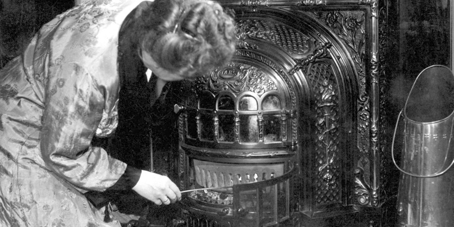 Vrouw in ochtendjas hurkt naast de rijkelijk versierde smeedijzeren haard en prikt met een pook in de brandende kolen, om het vuur op te stoken. Op de kachel een waterketel en ernaast een koperen kolenkit. 1947 - 1952 Vrouw in ochtendjas hurkt naast de rijkelijk versierde smeedijzeren haard en prikt met een pook in de brandende kolen, om het vuur op te stoken. Op de kachel een waterketel en ernaast een koperen kolenkit. 1947 - 1952