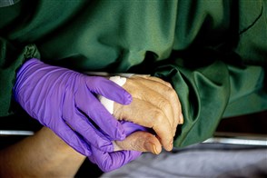 A nurse wearing purple gloves holds the hand of a patient.