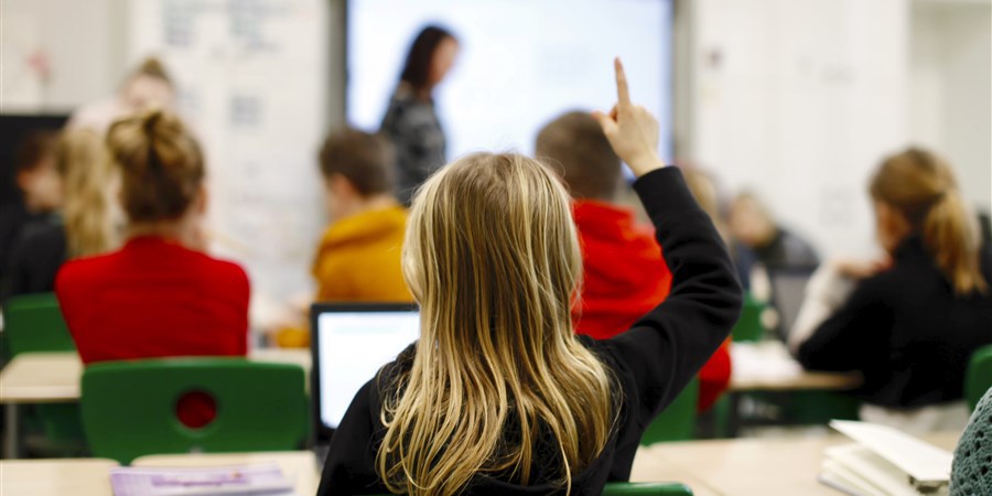 Kind  achterin klas. Steekt de vinger op. Op de achtergrond de juf.