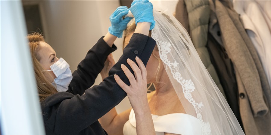 Bruid krijgt sluier omgeslagen door vrouw die zichzelf beschermt met handschoenen en een mondkapje.