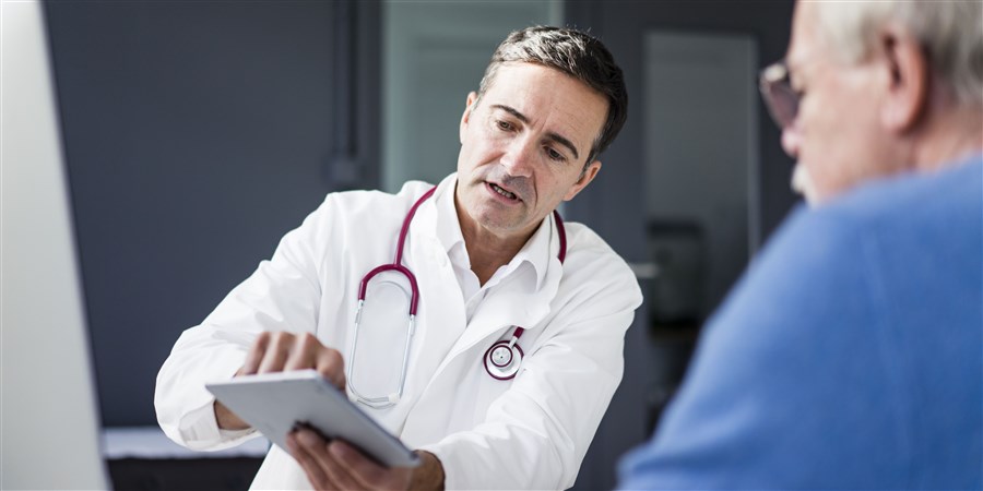 Dokter laat iets zien op tablet aan een patiënt, een oudere man met een bril en een blauwe trui