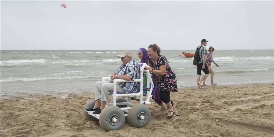 Twee vrouwen duwen oudere man in speciale rolstoel door het zand aan het strand.