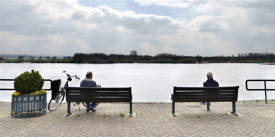 Twee oudere mannen rusten uit op een bankje aan de Waal, Rijn en kijken naar de schepen die voorbijkomen.
