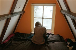 Jongere alleen, zittend op bed voor een beslagen raam op zolderkamer.