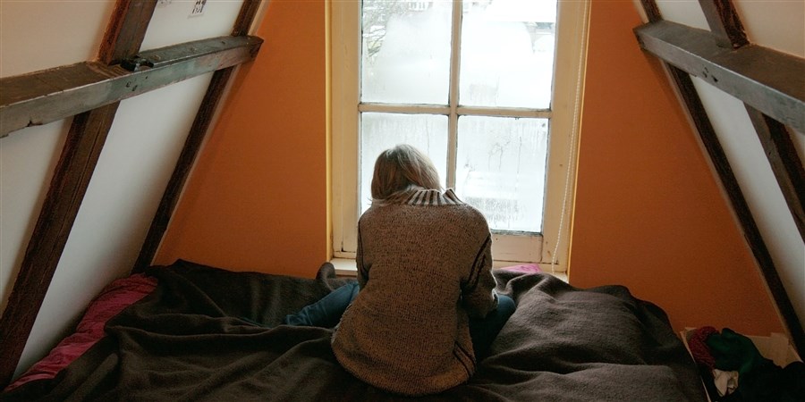 Jongere alleen, zittend op bed voor een beslagen raam op zolderkamer.